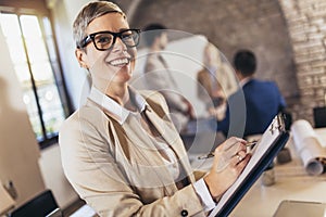 Young businesswoman writing on clipboard with team discussing project in the background