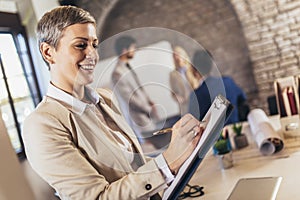 Young businesswoman writing on clipboard with team discussing project in the background