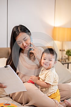 Young busy Asian mother is talking on the phone while taking care of her baby boy in bed