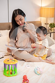 Young busy Asian mother is talking on the phone while taking care of her baby boy in bed