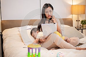 Young busy Asian mother is talking on the phone while taking care of her baby boy in bed