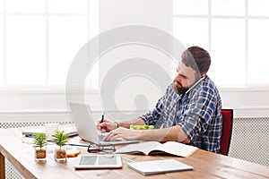 Young busy businessman with laptop has lunch in modern white office