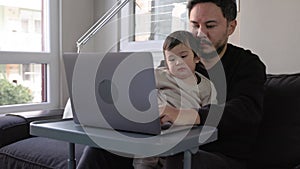 A young busy man working on laptop holding his son at home. Remote work