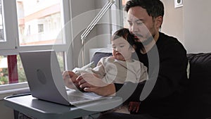 A young busy man working on laptop holding his son at home. Remote work