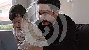 A young busy man working on laptop holding his son at home. Remote work