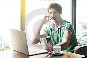 Young busy multiple businessman in green t-shirt sitting and working on laptop. talking on phone and looking at another smartphone