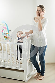 Young busy woman talking by phone while feeding her baby son with milk