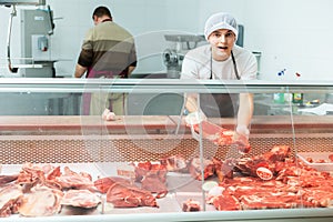 Young butcher shop seller arranging fresh beef in glass refrigerated display