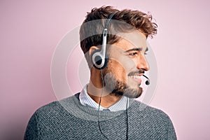 Young call center agent man with beard wearing headset over isolated pink background looking away to side with smile on face,