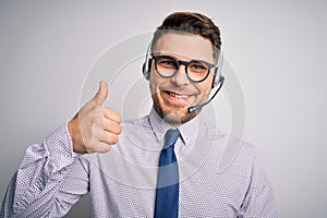 Young call center operator business man with blue eyes wearing glasses and headset doing happy thumbs up gesture with hand