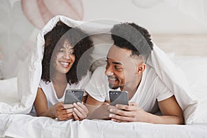 Young calm african loving couple girl guy in t-shirts using mobile cell phone lying on bed with white sheet pillow