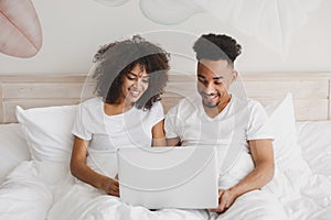 Young calm african loving couple girl guy in white t-shirts using laptop pc computer lying on bed indoors in bedroom at