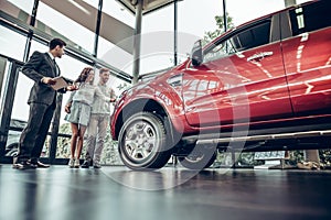 Young car salesman standing at the dealership telling about the features of the car to the customers