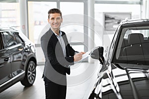 Young car salesman standing near new auto, taking notes and smiling at camera in automobile dealership, copy space