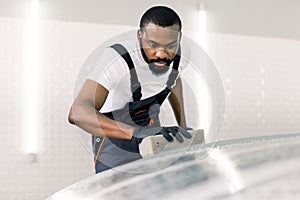 Young car service African male worker in black rubber gloves amd gray overalls, doing cleaning the hood of the car with