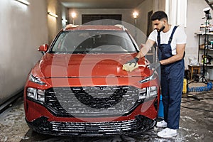 Young car service bearded male worker in black rubber gloves and blue overalls, cleaning the hood