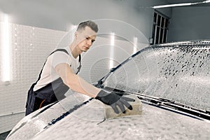 Young car service male worker in black rubber gloves doing cleaning the hood of the car with the help of shampoo and