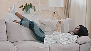 Young carefree african american woman lazily lying on sofa in room resting relaxing holding mobile phone chatting on