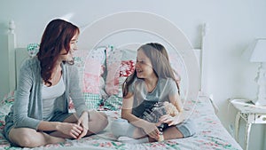 Young careful mother and little cheerful daughter talking and sharing secrets while sitting on bed at home in bedroom