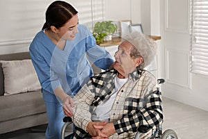 Young caregiver assisting senior woman in wheelchair indoors. Home health care service