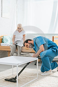 young caregiver cleaning carpet with vacuum cleaner while senior woman