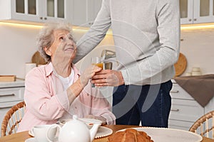 Young caregiver giving drink to senior woman in kitchen. Home care service