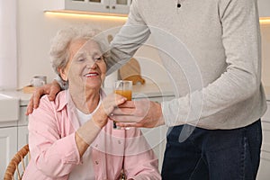 Young caregiver giving drink to senior woman in kitchen. Home care service
