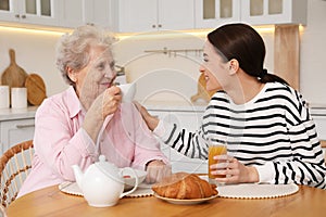 Young caregiver and senior woman having breakfast at table in kitchen. Home care service