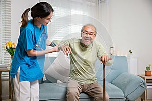 Young carer supporting helping senior man stand up with walking stick from sofa