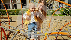 Young caring mother helping her baby son climbing up on the rope net at playground. Children playing outdoor, kids outside, summer