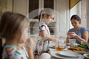 Young caring mother and her two little daughters eating pancakes with honey at the breakfast in the cozy kitchen