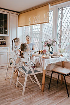 Young caring mother and her two little daughters have a breakfast in the light kitchen with large window