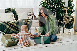 Young caring mother with toddler open Christmas gifts while sitting on the floor at home.