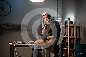 A young carpenter writes a message on his smartphone in a carpenter`s workshop while sitting on his Desk at lunchtime.