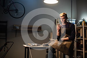 A young carpenter writes a message on his smartphone in a carpenter`s workshop while sitting on his Desk at lunchtime.