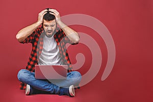 Young casual business man sitting on the floor with a laptop surprised and shocked isolated over red background
