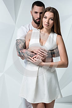 Young casual couple with woman holding his hands over woman`s shoulder and looking for the camera. On white background