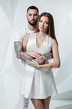 Young casual couple with woman holding his hands over woman`s shoulder and looking for the camera. On white background