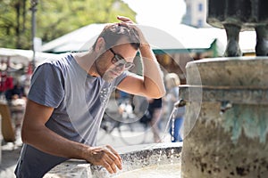 Young casual cucasian man refreshing himself with water from public city fountain on a hot summer day