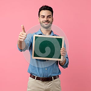 Young casual guy making thumbs up sign and holding blackboard