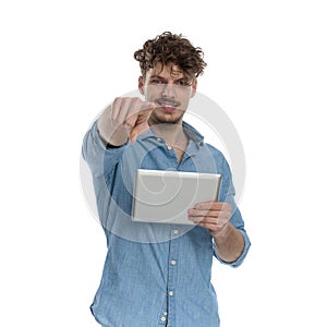 Young casual guy smiling, holding tab and pointing finger