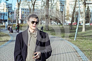 Young casual smoker man with sunglasses in the black coat smoking cigarette outside in the park