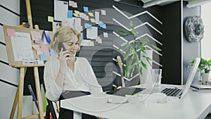 Young casual woman sitting at desk using laptop, answering call, talking on the phone
