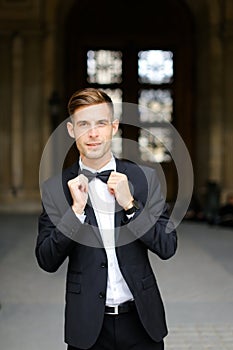 Young caucaisian man standing and posing, wearing black suit and bow tie.