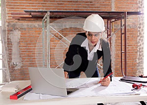 Young caucasian architect woman looking laptop and working drawing blueprint for planning building house on desk.
