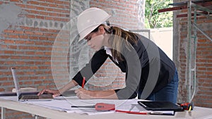 Young caucasian architect woman looking laptop and working drawing blueprint for planning building house on desk.