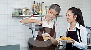 Young caucasian barista pouring milk from the jug into a paper coffee cup. Asian assistant in an apron holding croissant plate