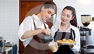 Young caucasian barista pouring milk from the jug into a paper coffee cup. Asian assistant in an apron holding croissant plate