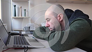 Young Caucasian bearded bald man is bored at his desk near laptop. Depressed person. Psychology and mental health