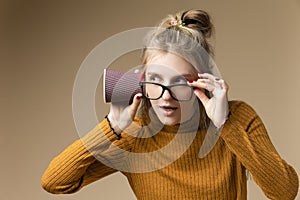 Young Caucasian Blond Girl Listening To Arcane Hidden Gossips and Secrets Using Disposable paper Drinking Cup Over Beige